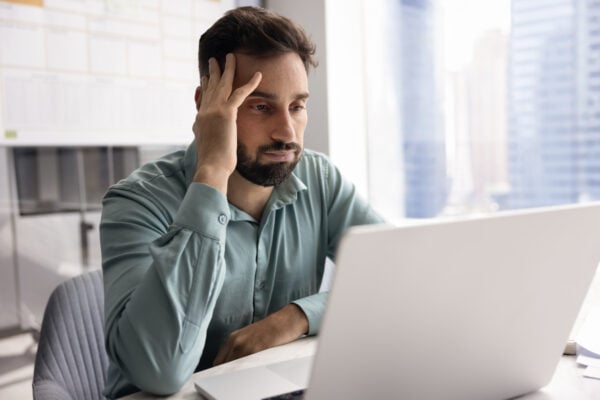A man sitting at a desk with his hand on his head, looking frustrated at a laptop screen.