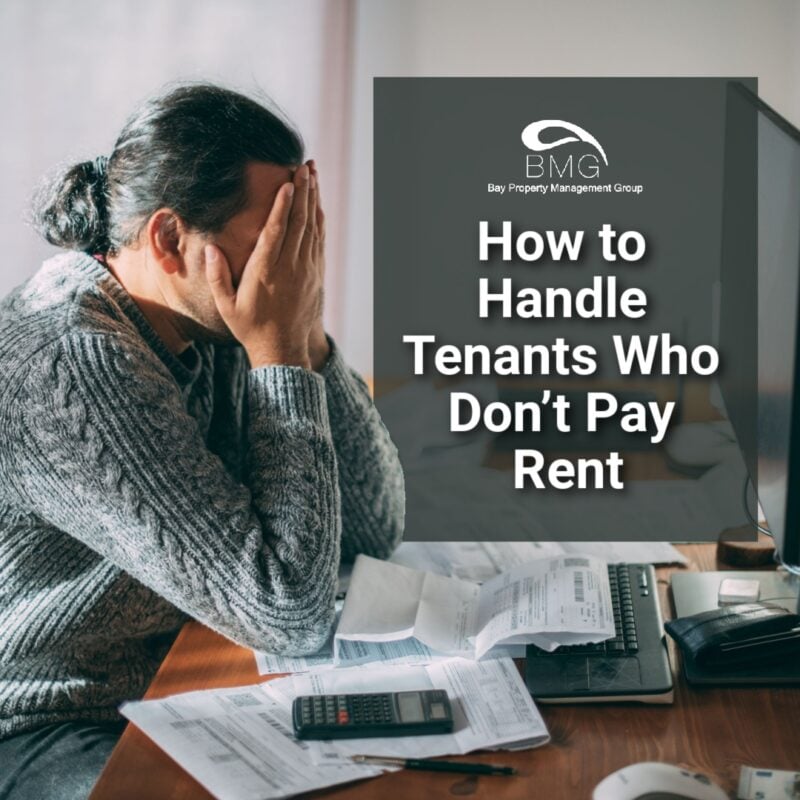 A stressed man is sitting at a desk with his head in his hands, surrounded by bills, documents, and a calculator. The text overlay reads: "How to Handle Tenants Who Don’t Pay Rent," along with the logo for Bay Property Management Group.